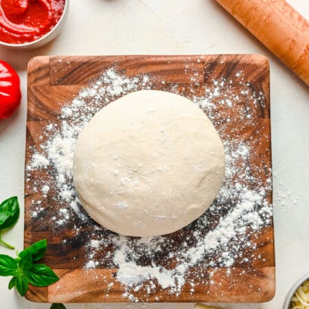 A ball of pizza dough on a wooden cutting board next to a dish of pizza sauce.