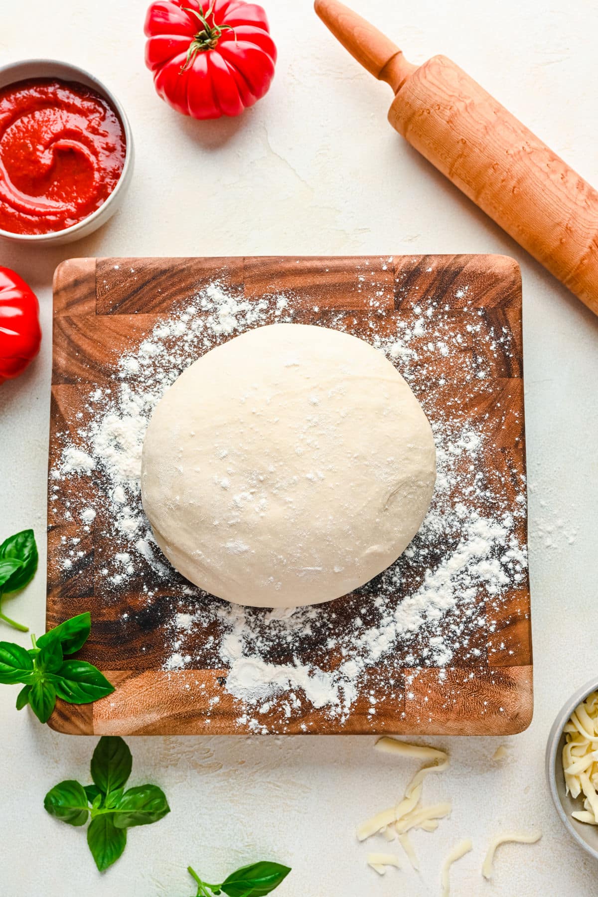 A ball of pizza dough on a wooden cutting board next to a dish of pizza sauce.