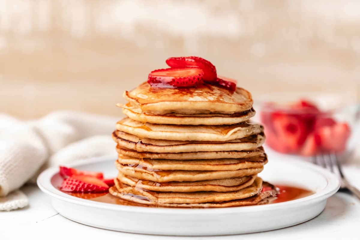 A white plate with a stack of oatmeal pancakes topped with strawberries next to a dish of strawberries.