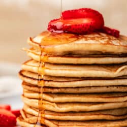 A stack of oatmeal pancakes with maple syrup pouring onto it topped with strawberries.