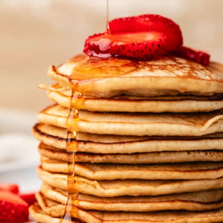 A stack of oatmeal pancakes with maple syrup pouring onto it topped with strawberries.
