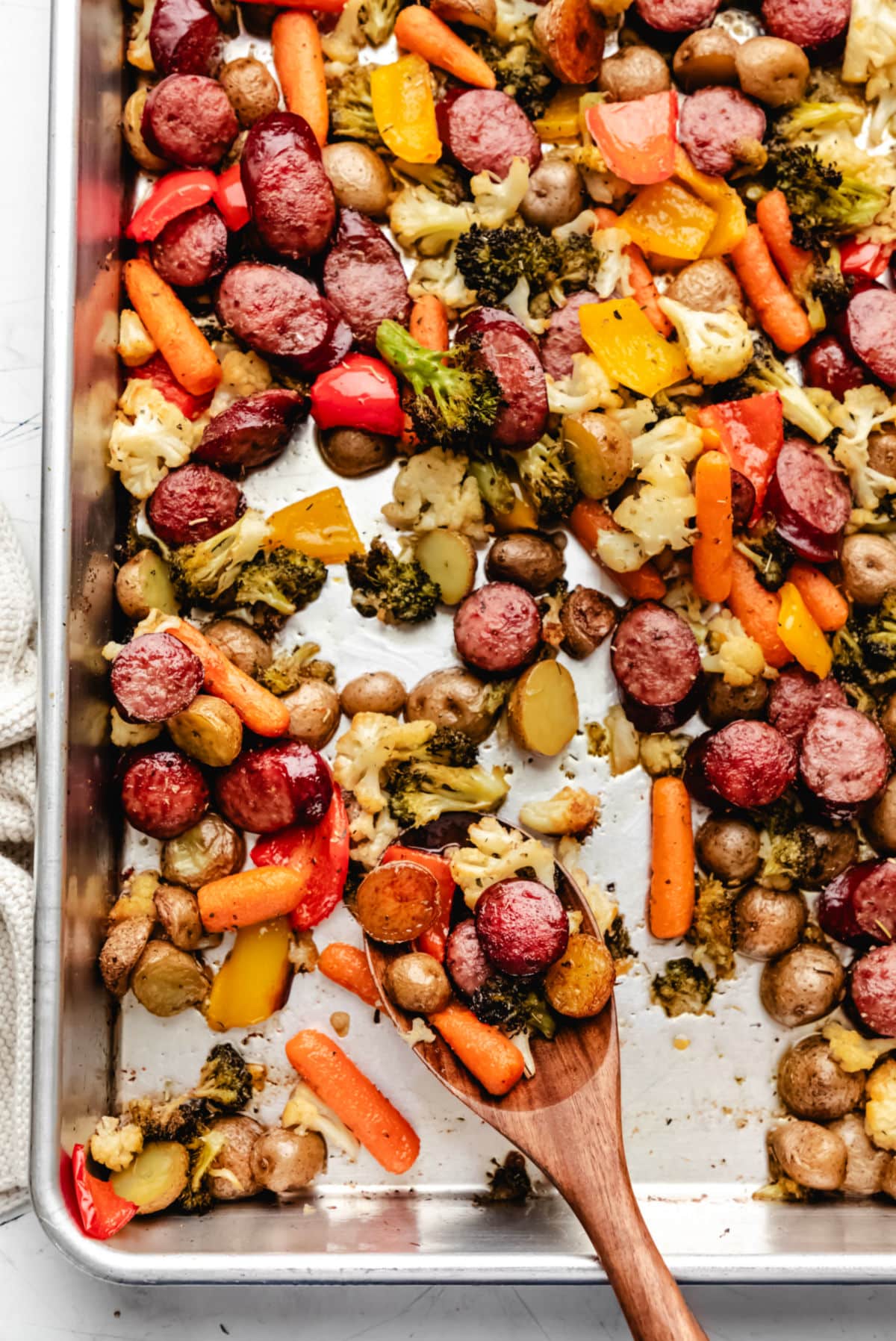A wooden spoon holding a scoop of sheet pan sausage and veggies. 