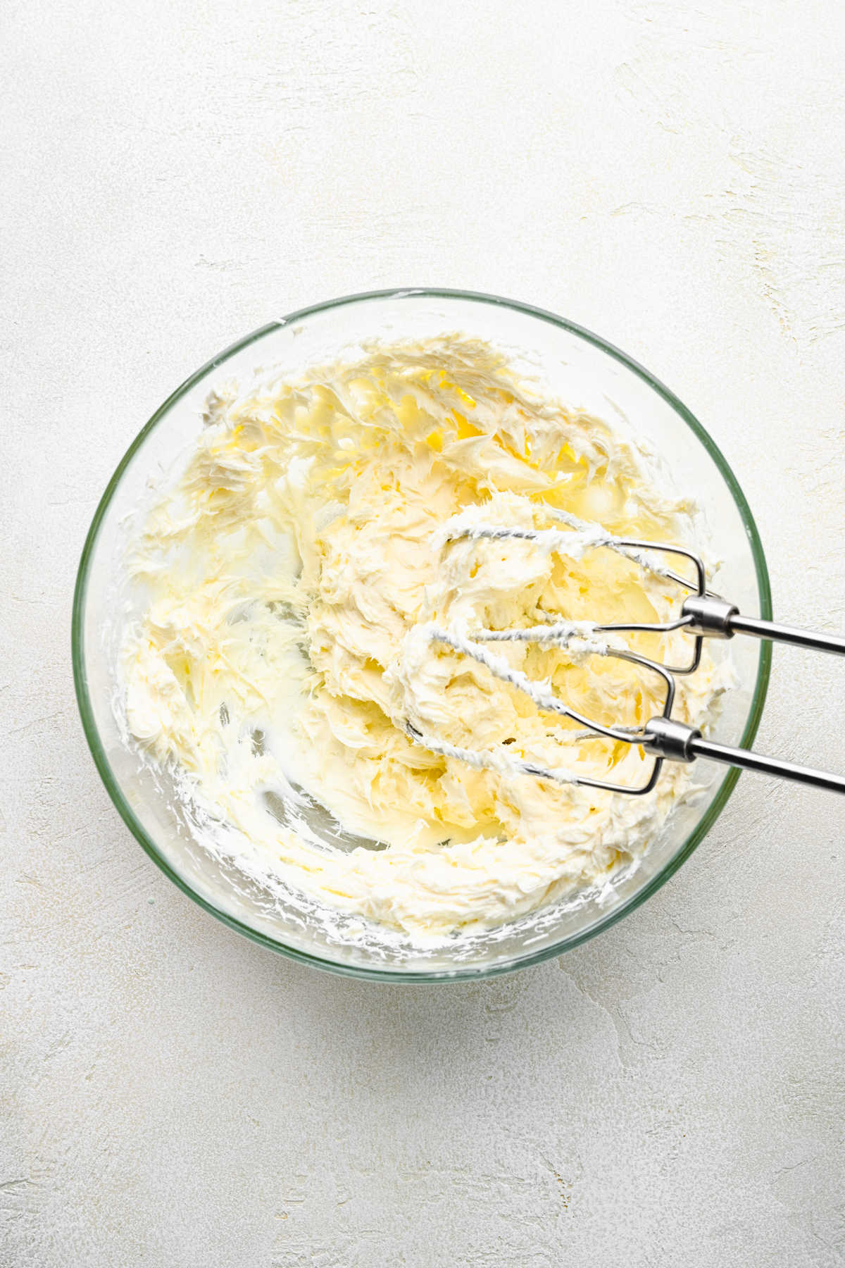 A hand mixer beating cream cheese and butter together in a glass mixing bowl. 