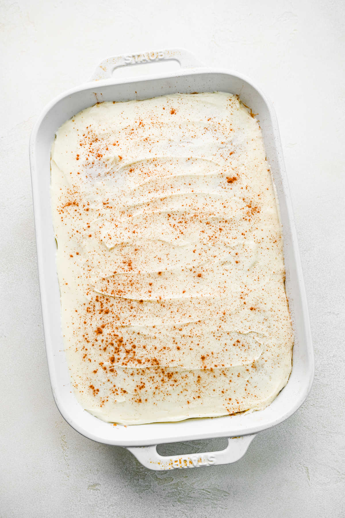 Frosted cinnamon roll poke cake in a white baking pan with cinnamon sprinkled on top.