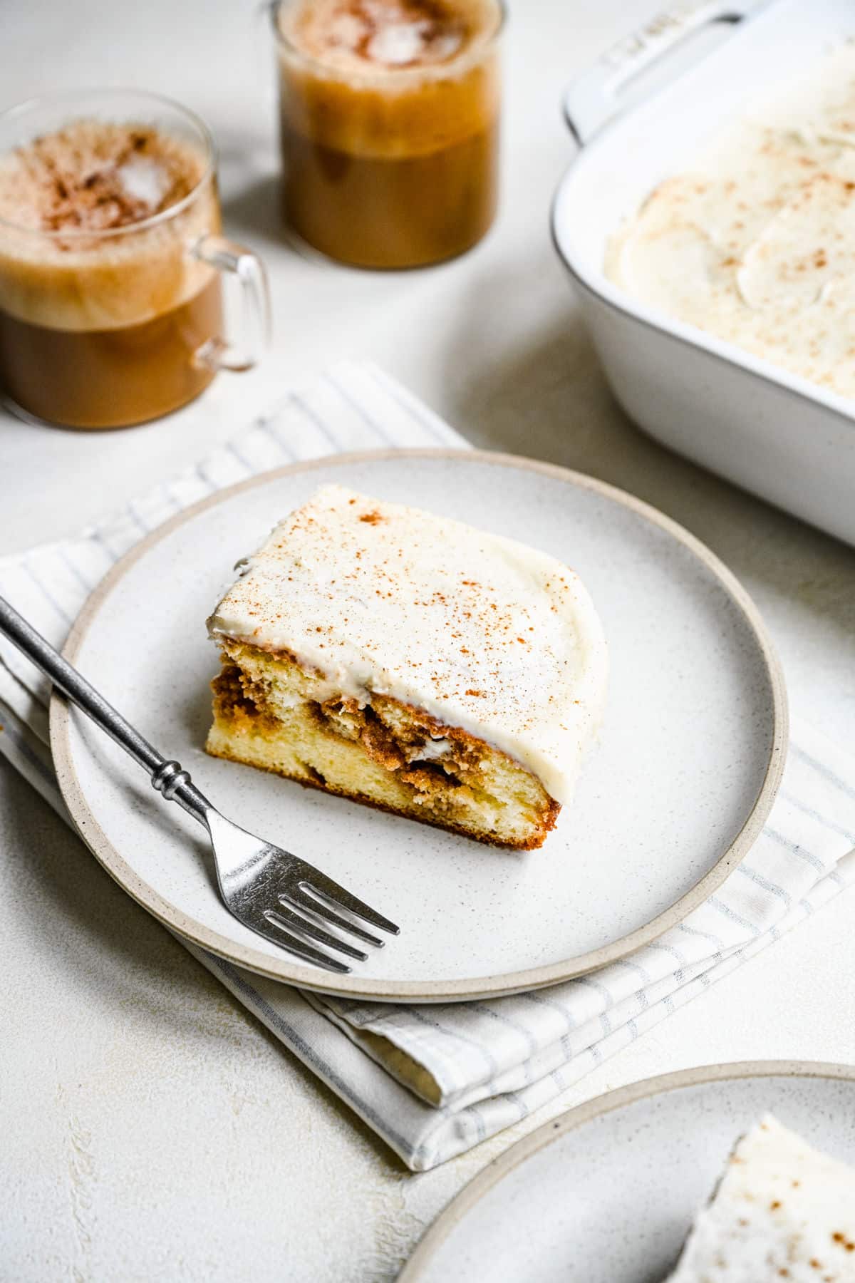 A slice of cinnamon roll poke cake on a white plate next to two cups of coffee.