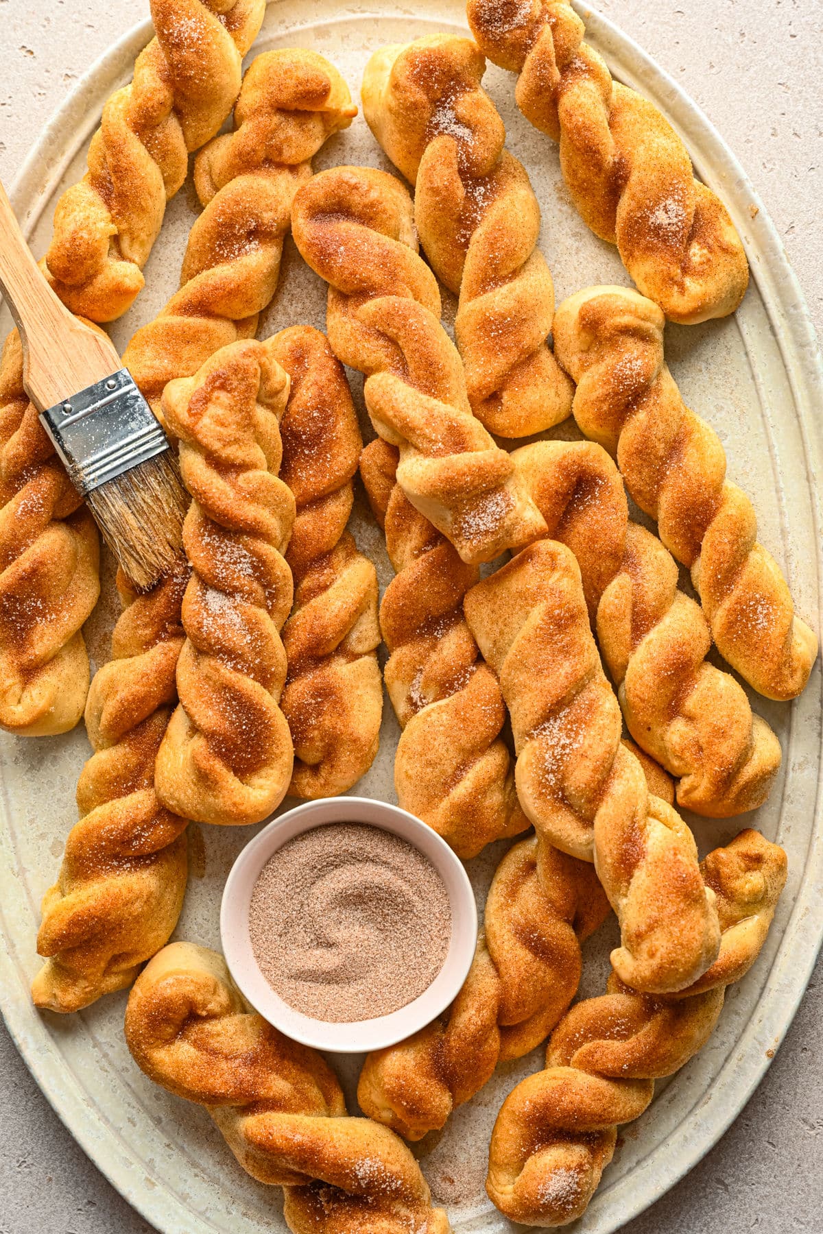 A platter full of stacked cinnamon breadsticks and a dish of cinnamon sugar. 