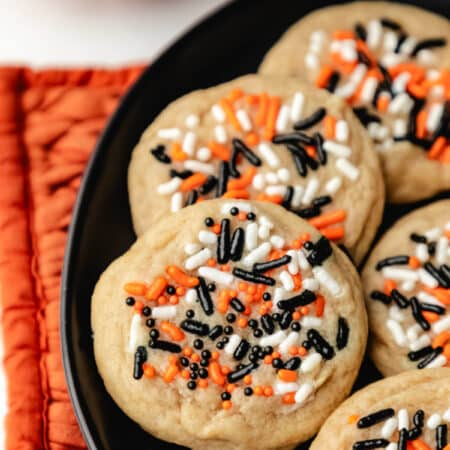 Halloween sprinkle cookies on a black plate next to a jack-o-lantern mug.
