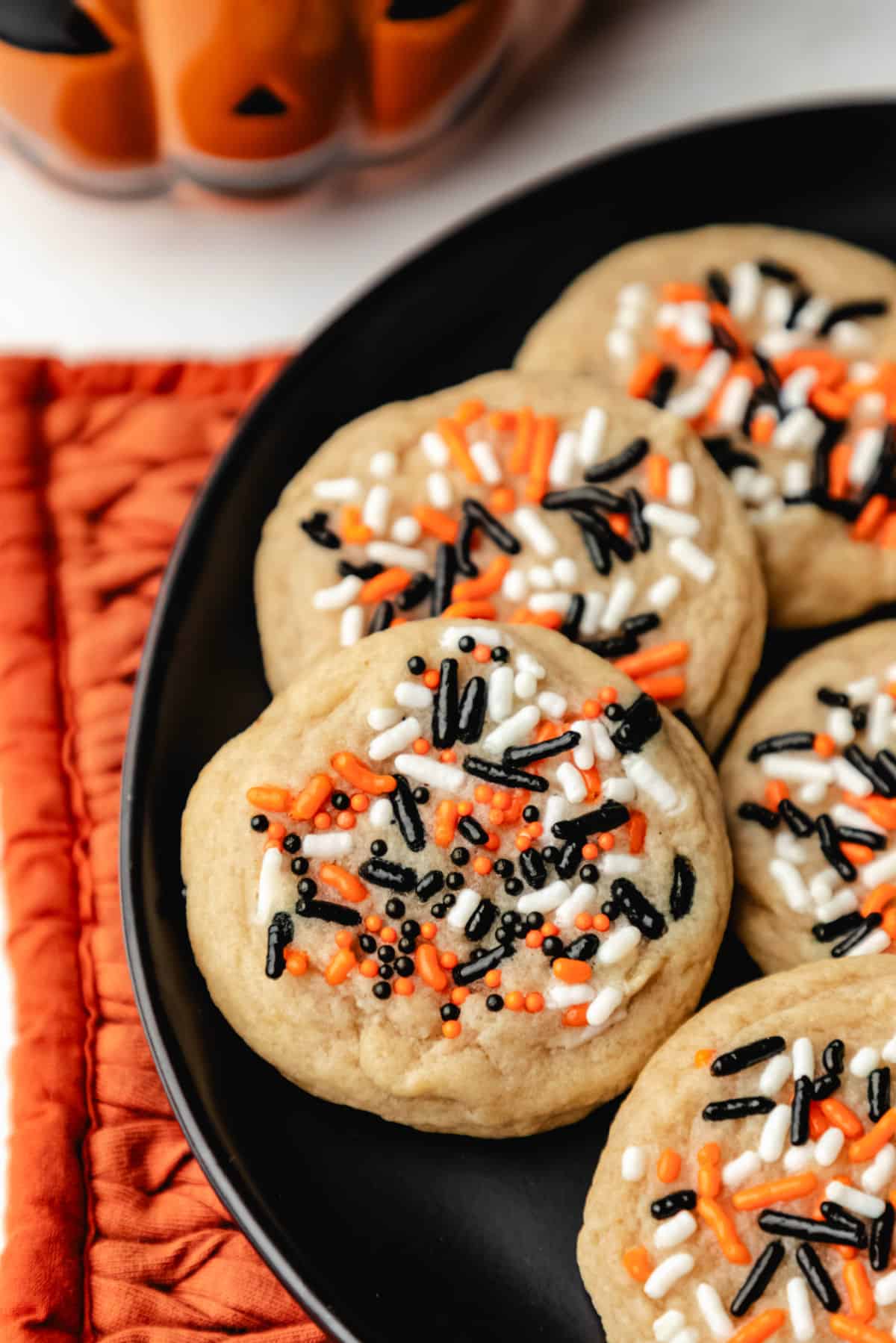 Halloween sprinkle cookies on a black plate next to a jack-o-lantern mug.