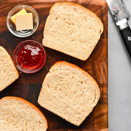 Slices of homemade white bread on a cutting board next to dishes of butter and jam.
