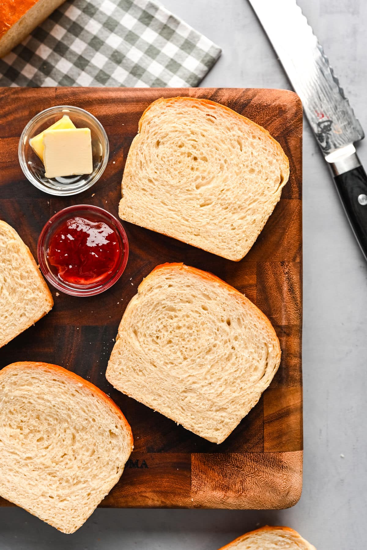 Slices of homemade white bread on a cutting board next to dishes of butter and jam.