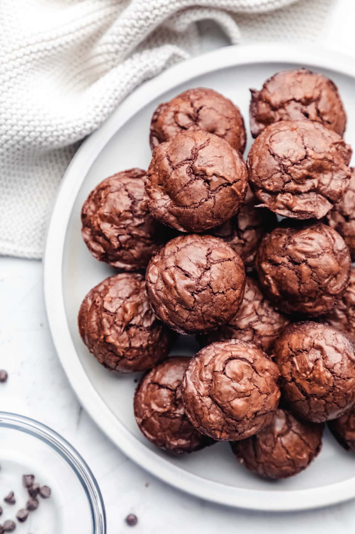 A white plate full of brownie bites next to a glass bowl of mini chocolate chips. 