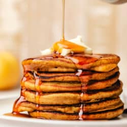 Maple syrup pouring out of a ceramic syrup holder onto a stack of pumpkin pancakes.