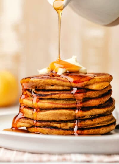 Maple syrup pouring out of a ceramic syrup holder onto a stack of pumpkin pancakes.