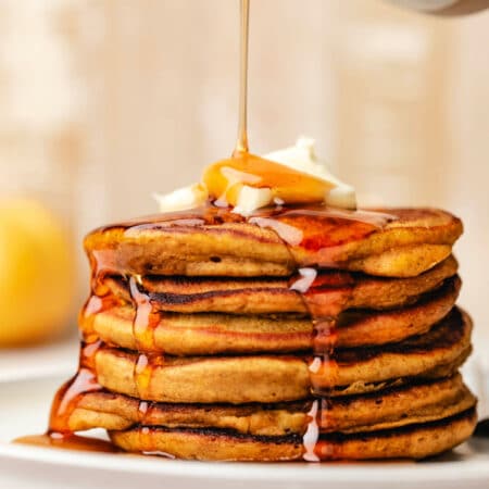 Maple syrup pouring out of a ceramic syrup holder onto a stack of pumpkin pancakes.