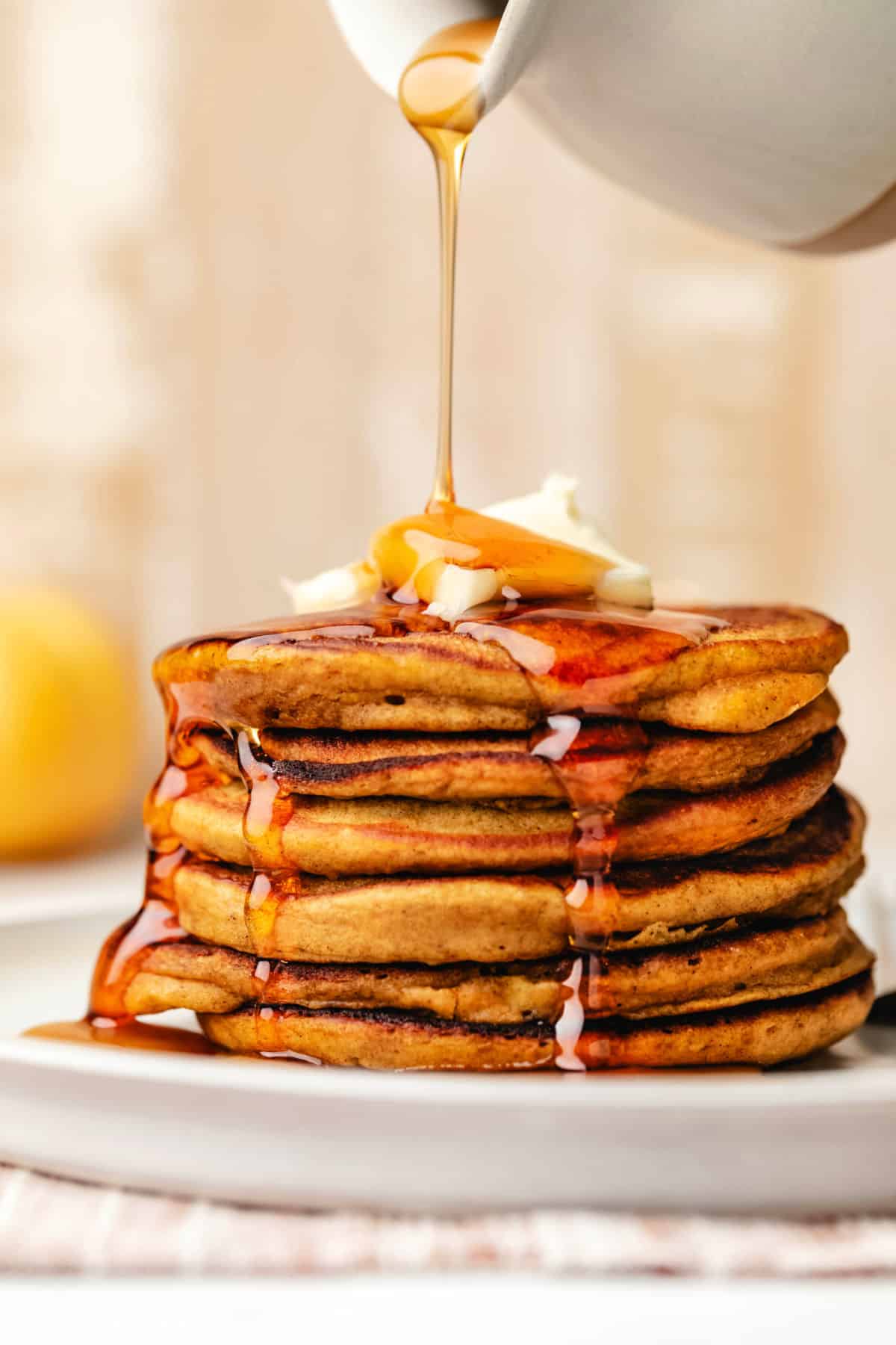 Maple syrup pouring out of a ceramic syrup holder onto a stack of pumpkin pancakes.