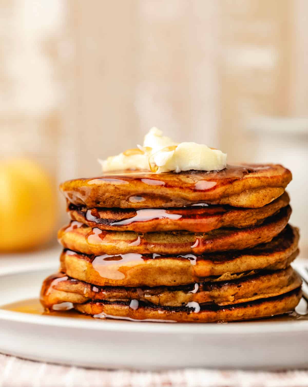 A stack of pumpkin pancakes topped with two pieces of butter and maple syrup running down the sides. 