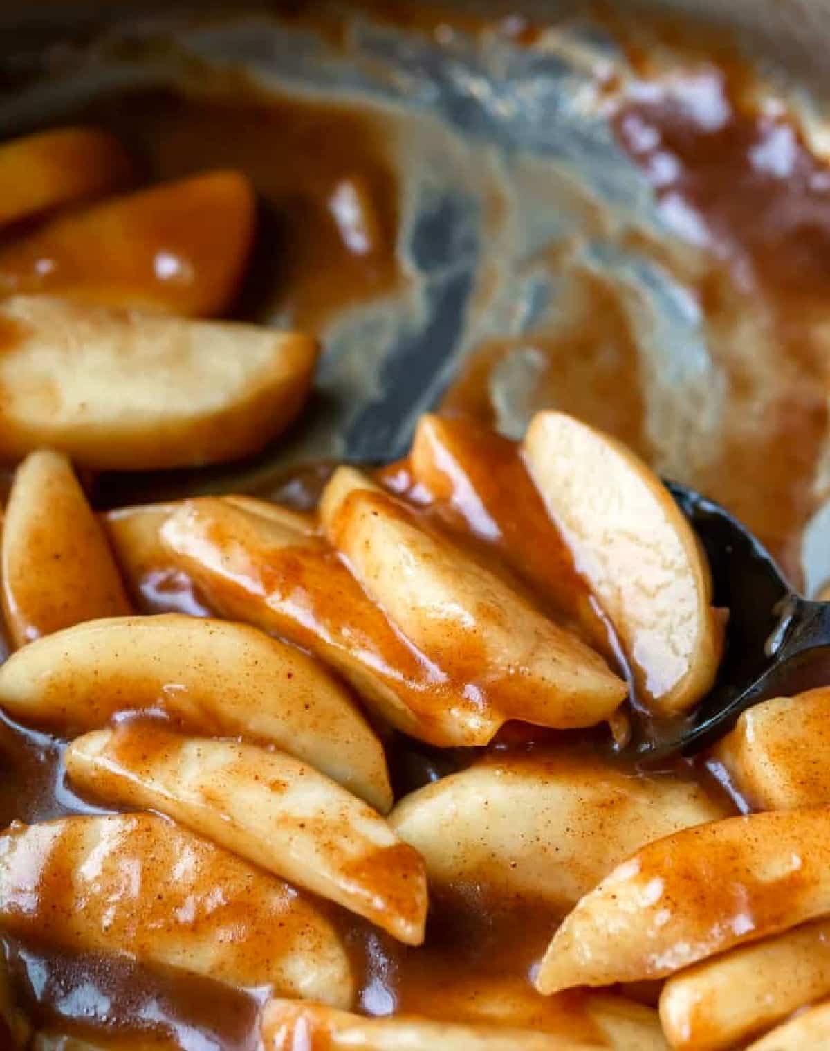 A black spoon holding a scoop of cinnamon apples in a silver skillet. 