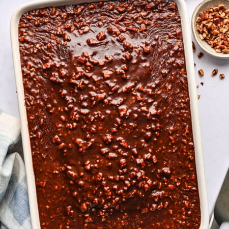 Frosted Coca Cola cake in a white pan next to dishes of cocoa powder and chopped nuts.