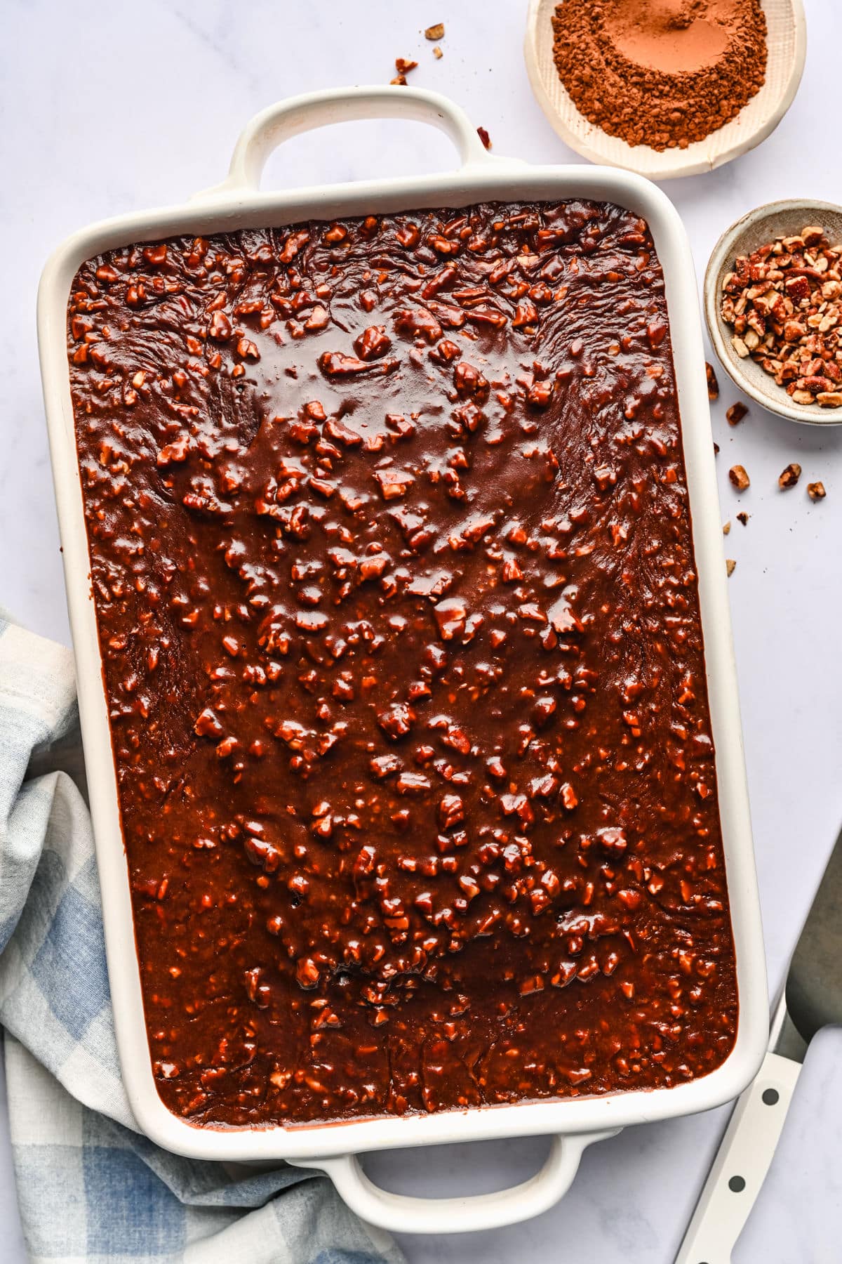 Frosted Coca Cola cake in a white pan next to dishes of cocoa powder and chopped nuts.