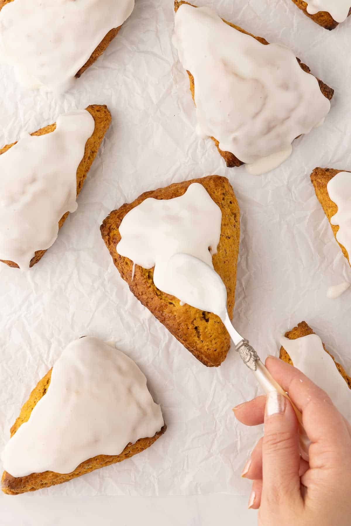 A spoon spreading white glaze onto the top of a pumpkin scone. 