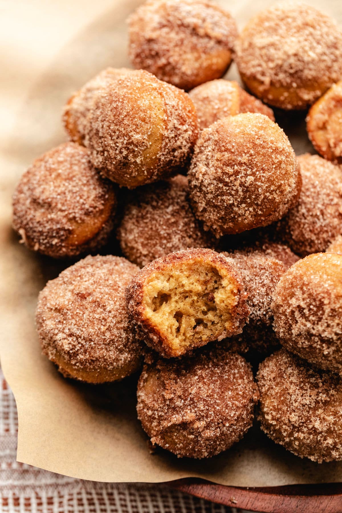 A wooden cake stand lined with brown parchment paper with a stack of baked apple cider donut holes on top.