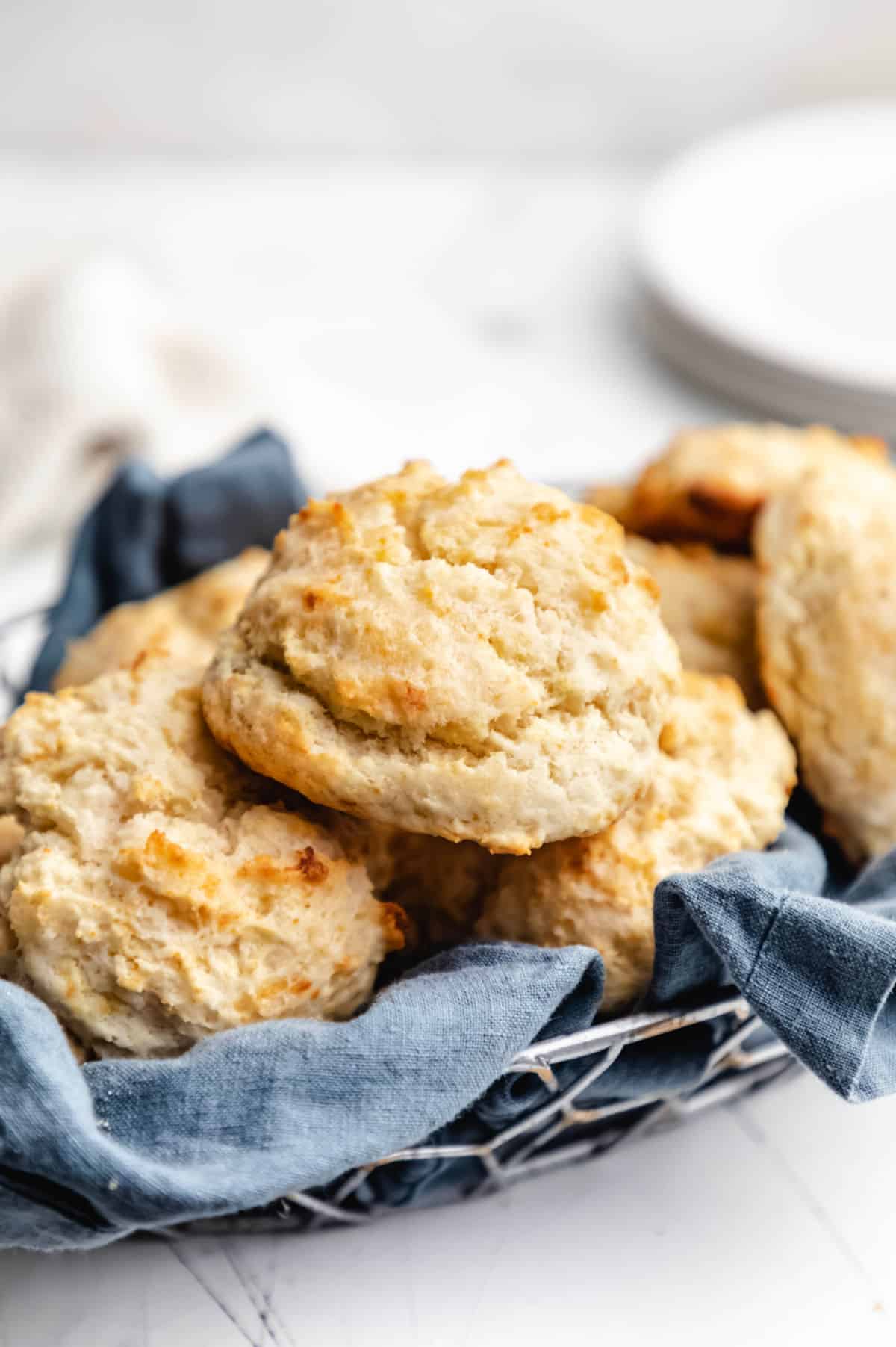 A basket of buttermilk drop biscuits with a stack of white plate next to it.
