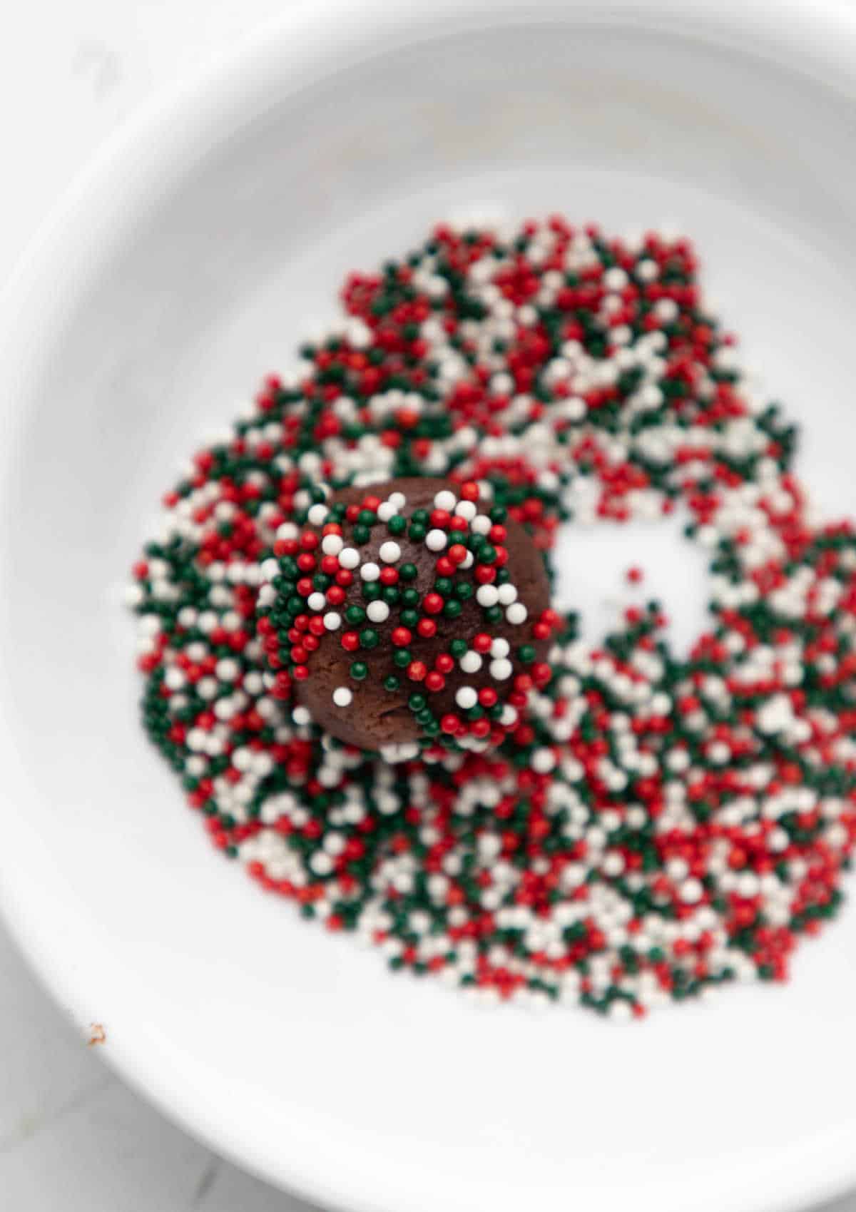 A ball of chocolate cookie dough rolling in a dish of sprinkles. 