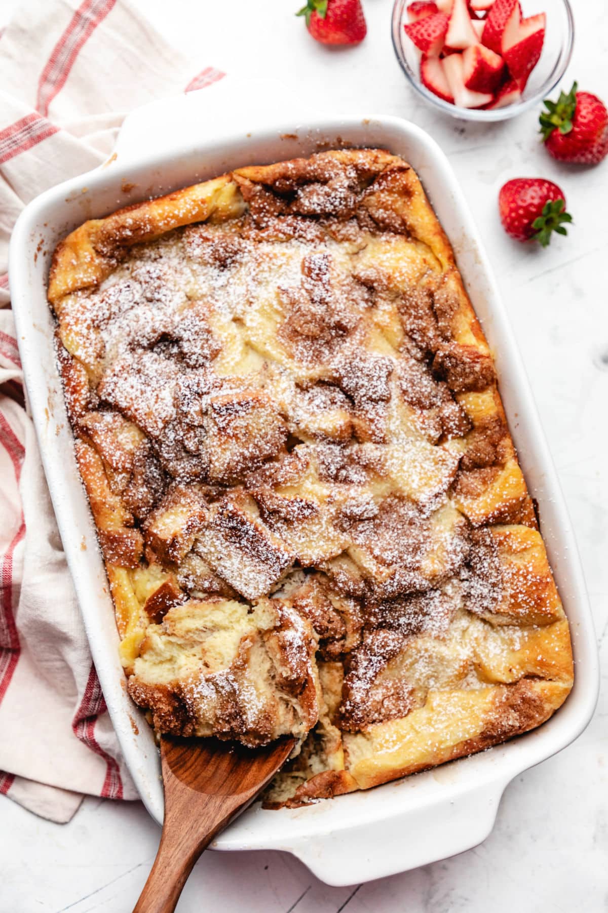 A wooden spoon holding a scoop of French toast casserole in a white baking pan.