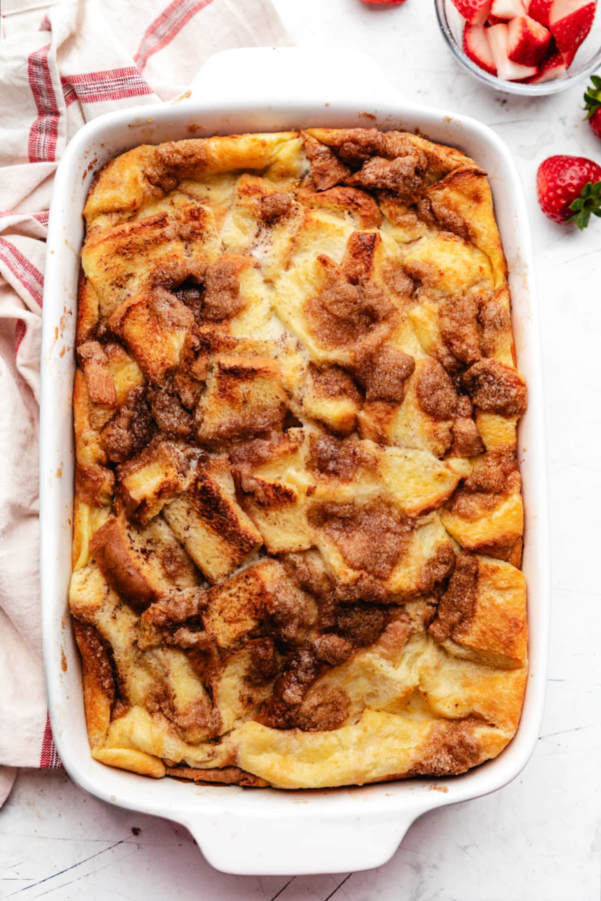 A baked French toast casserole in a white baking dish next to a bowl of sliced strawberries.