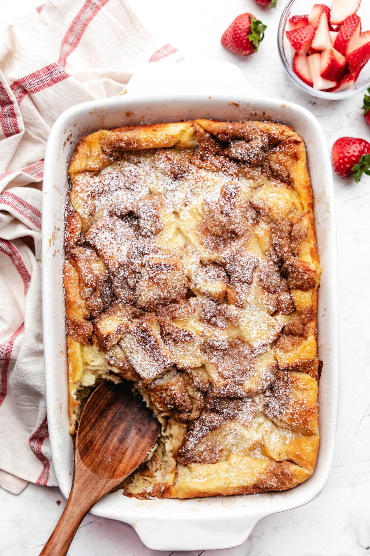 A white baking dish of overnight French toast casserole next to a striped kitchen towel. 
