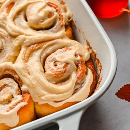 A white baking dish full of maple cinnamon rolls next to a glass dish of maple syrup.