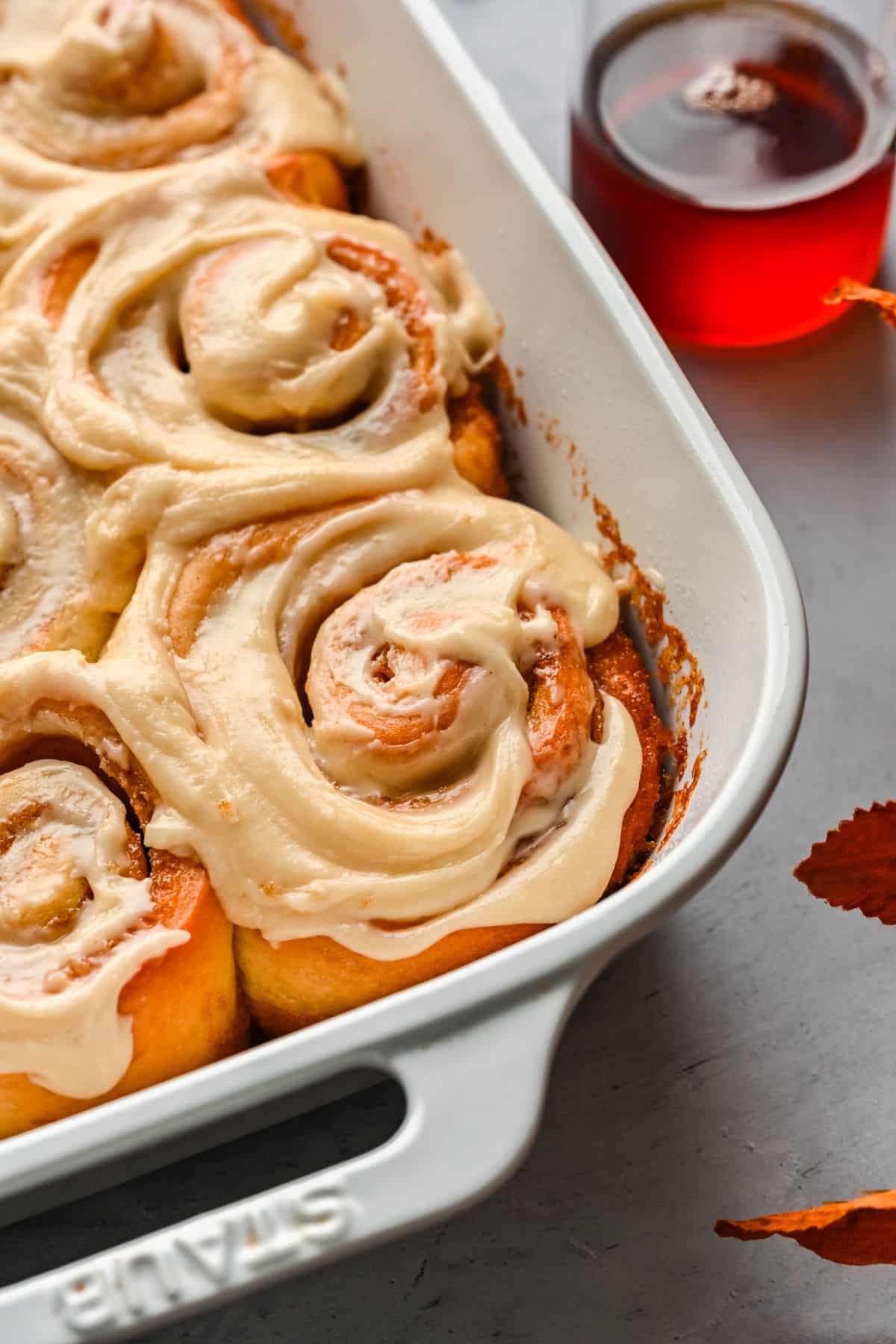 A white baking dish full of maple cinnamon rolls next to a glass dish of maple syrup. 