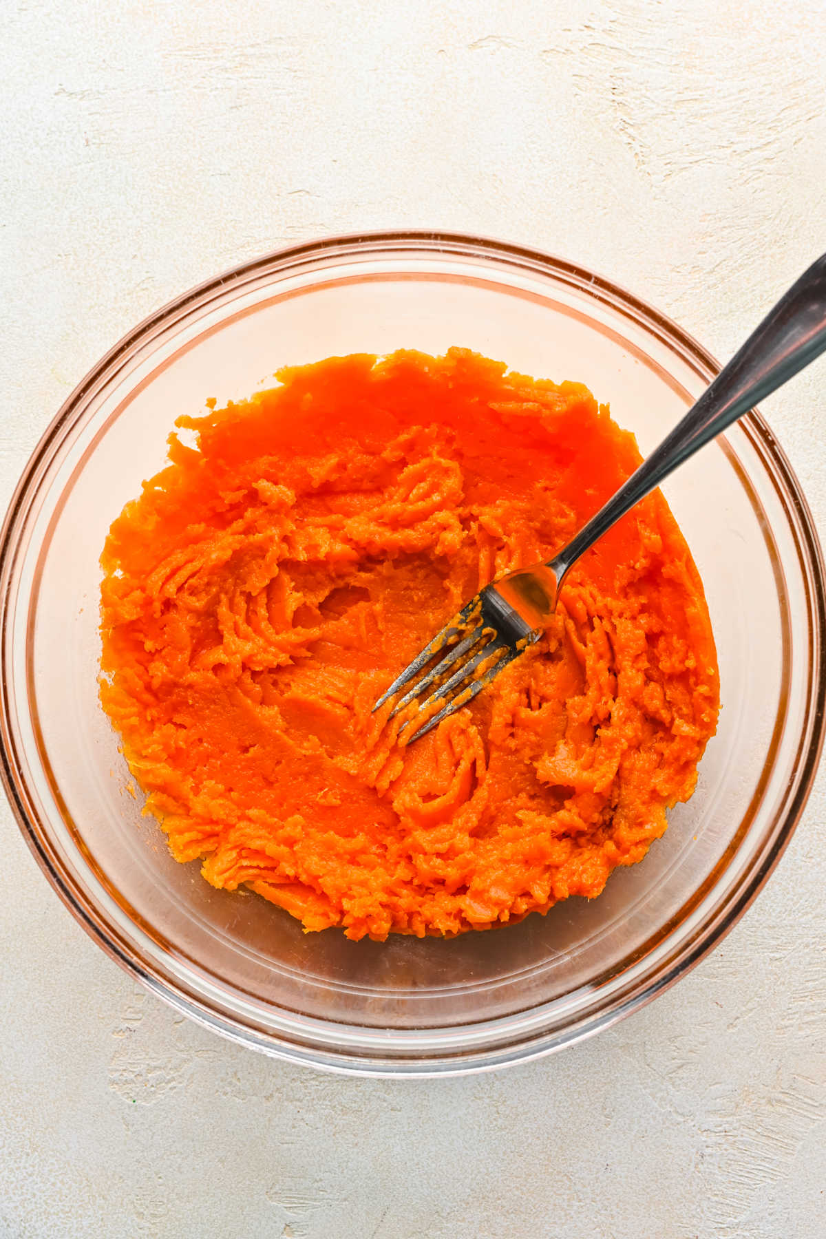 A fork mashing sweet potato in a glass mixing bowl. 