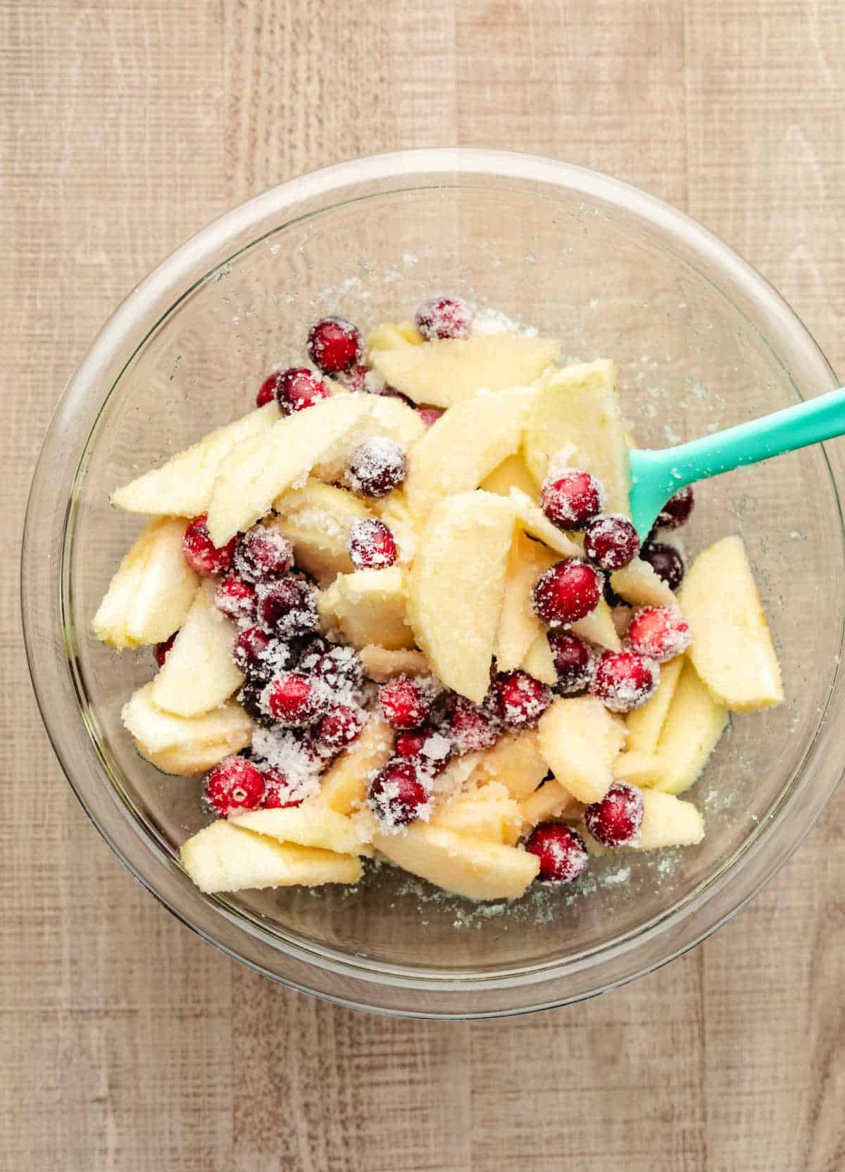 A glass mixing bowl with apple slices and cranberries coated in sugar. 