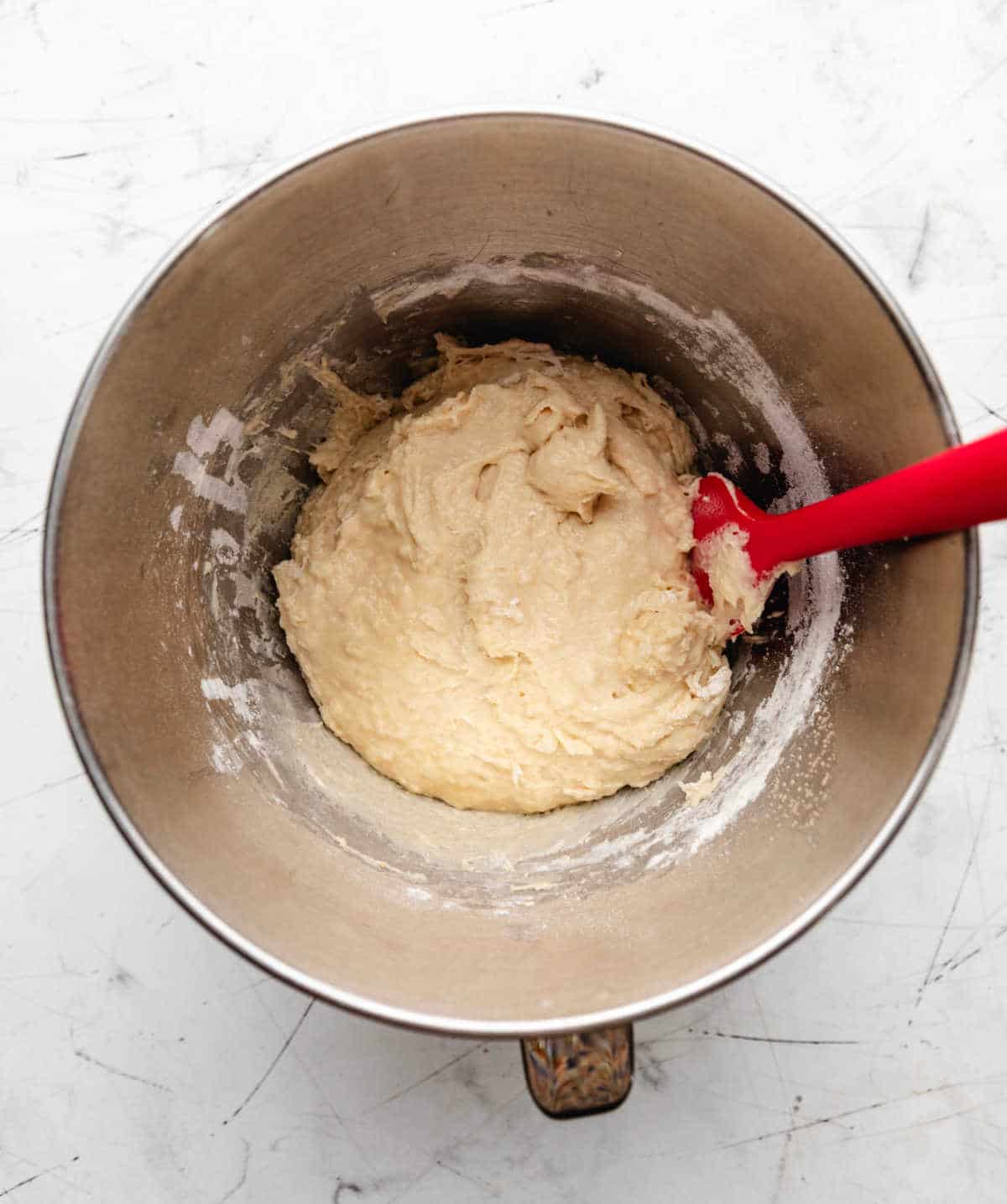 A red spatula mixing potato roll dough in a silver mixing bowl. 