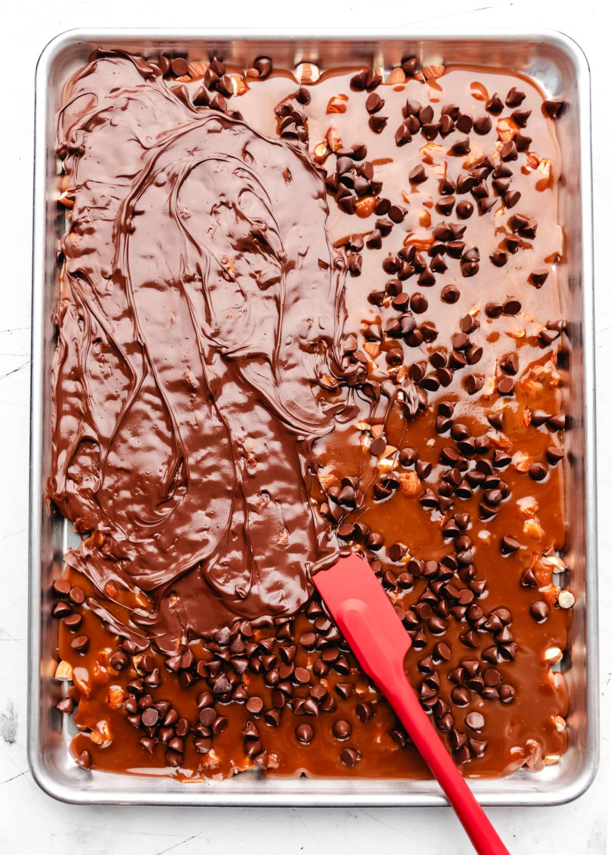 A red silicone spatula spreading melted chocolate chips over toffee.