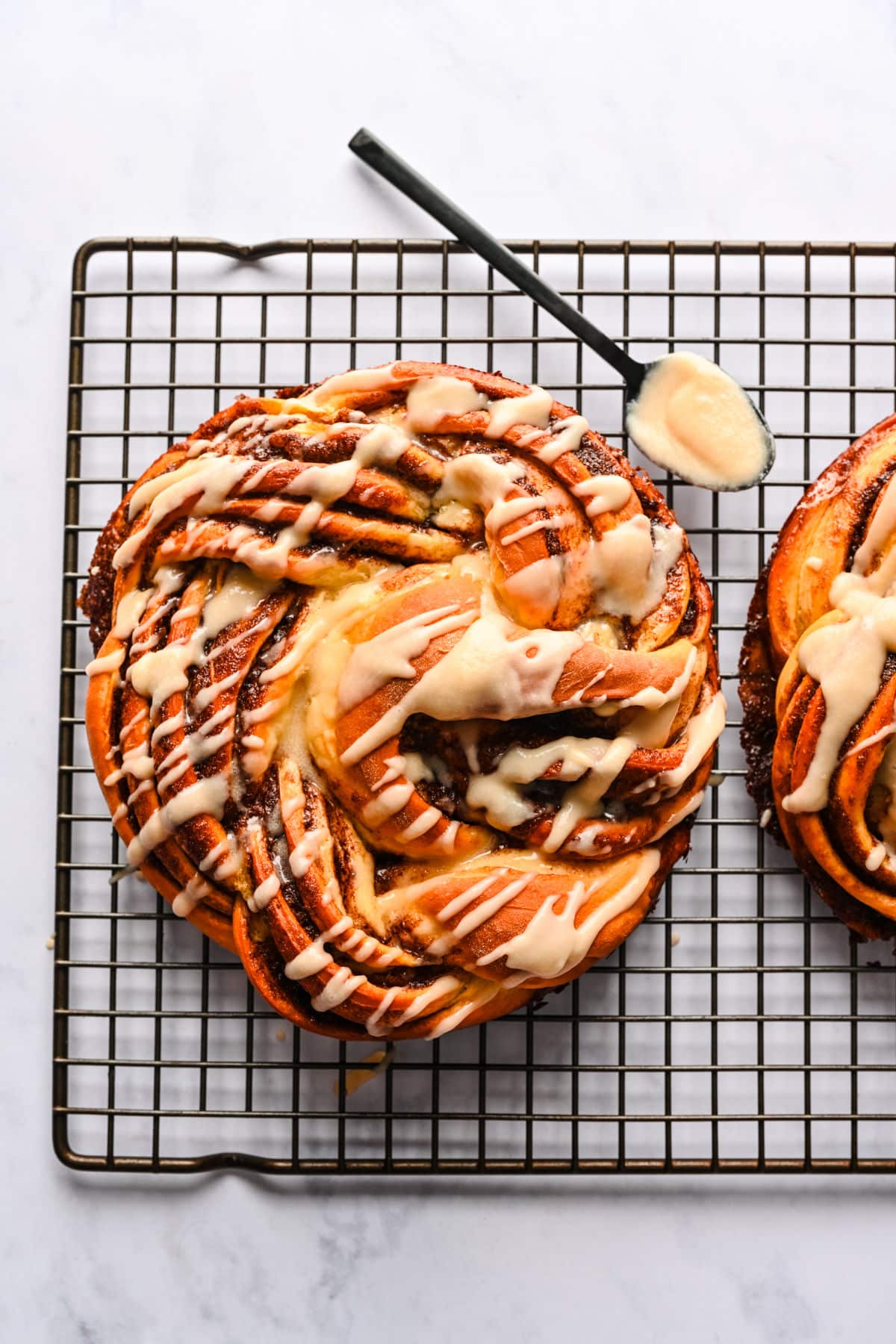 A loaf of braided cinnamon bread on a wire rack next to a spoon full of icing.