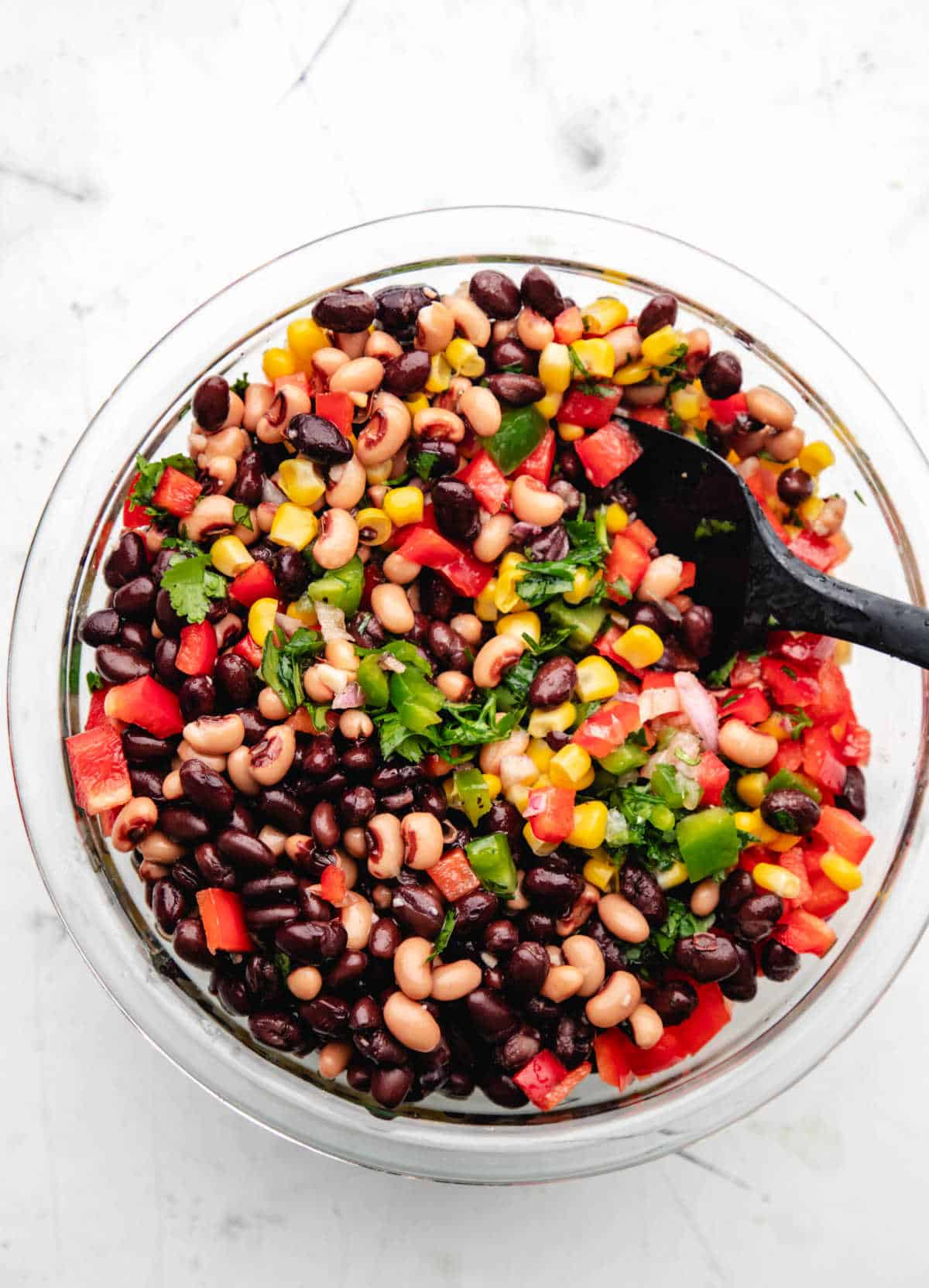 A black spoon stirring up cowboy caviar in a glass mixing bowl.
