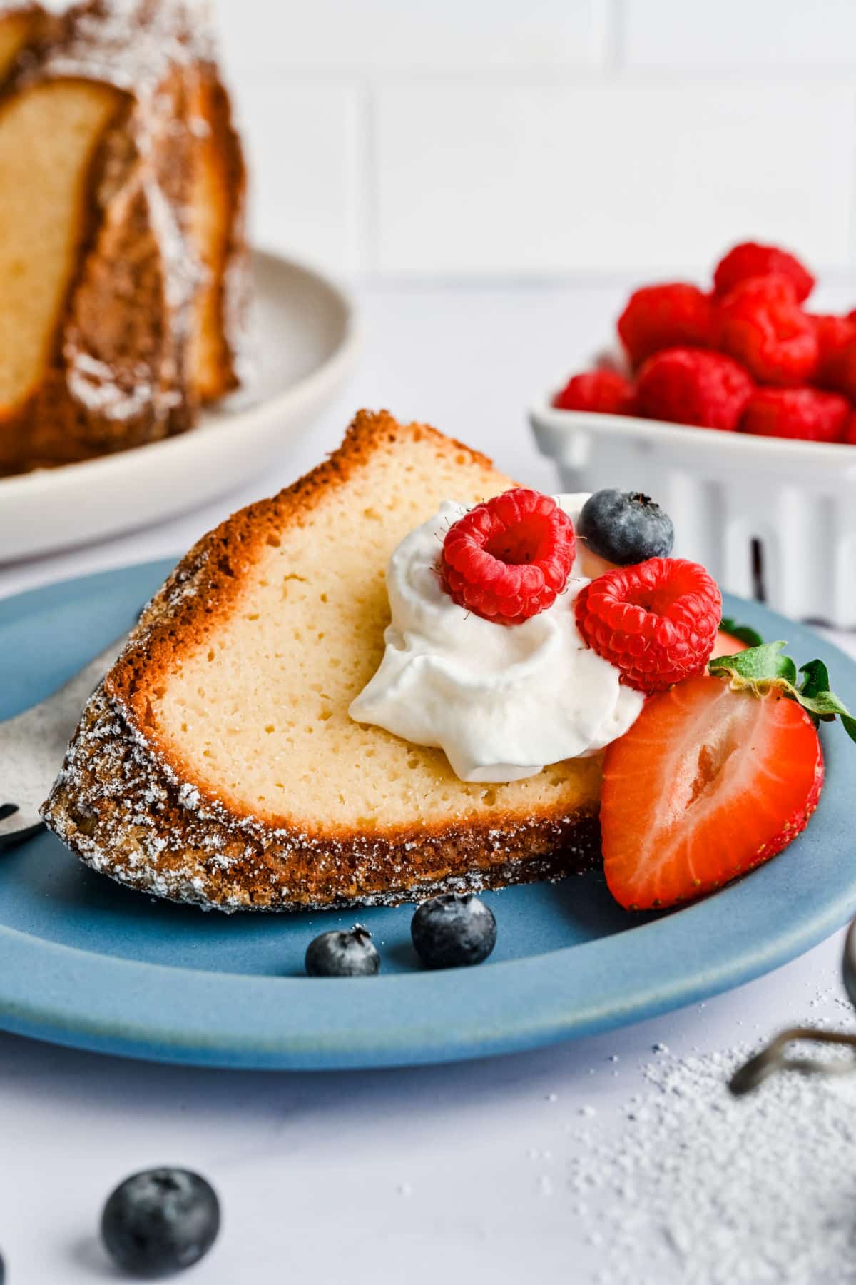 A slice of Greek yogurt pound cake on a blue plate next to a bowl of raspberries. 