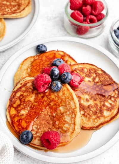 A plate of cottage cheese pancakes next to bowls of raspberries and blueberries.