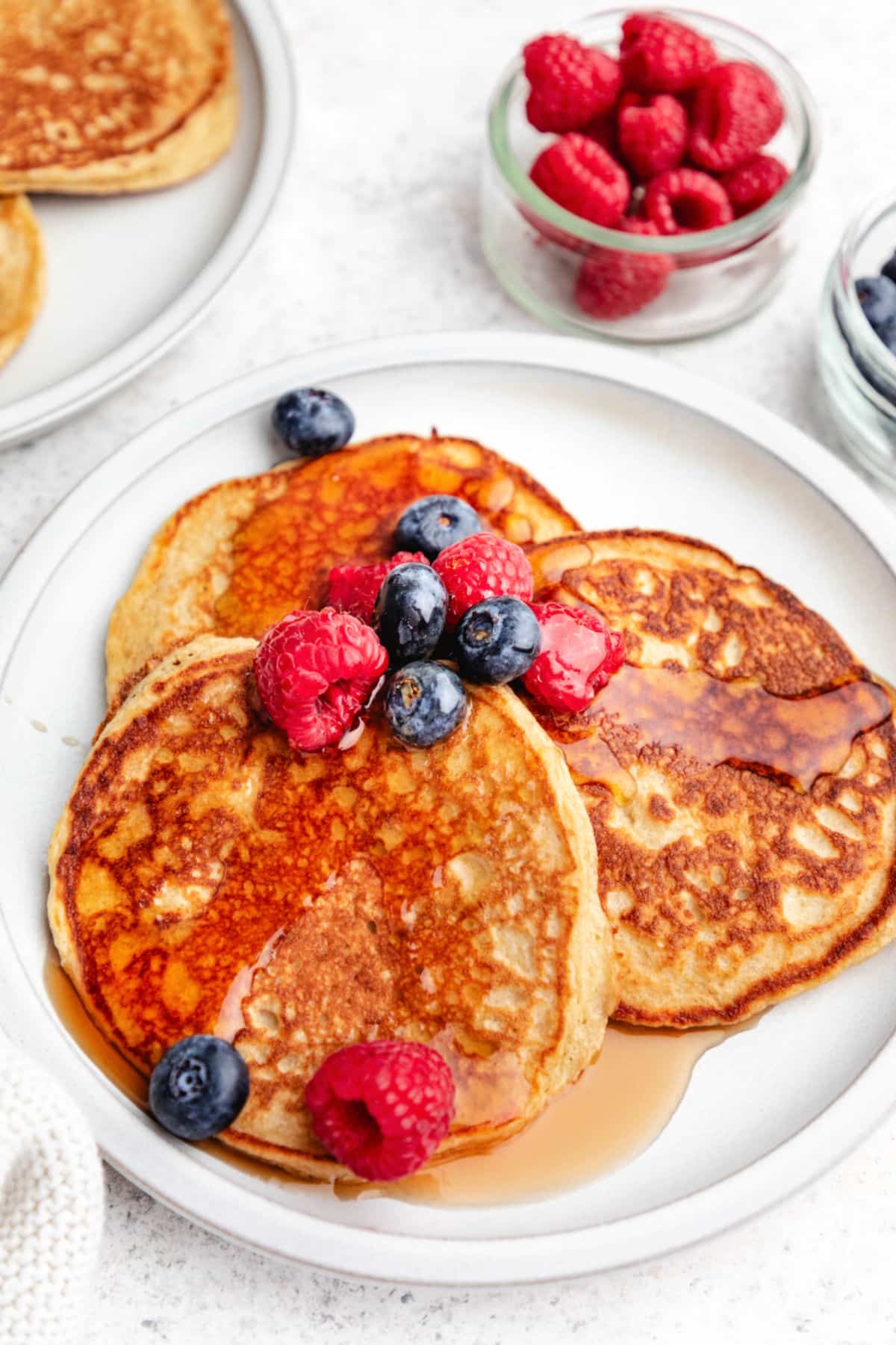 A plate of cottage cheese pancakes next to bowls of raspberries and blueberries.