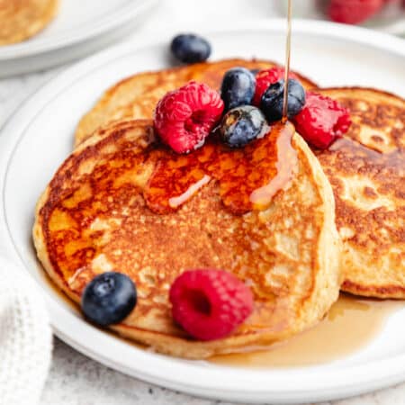 Maple syrup pouring onto cottage cheese pancakes topped with berries.