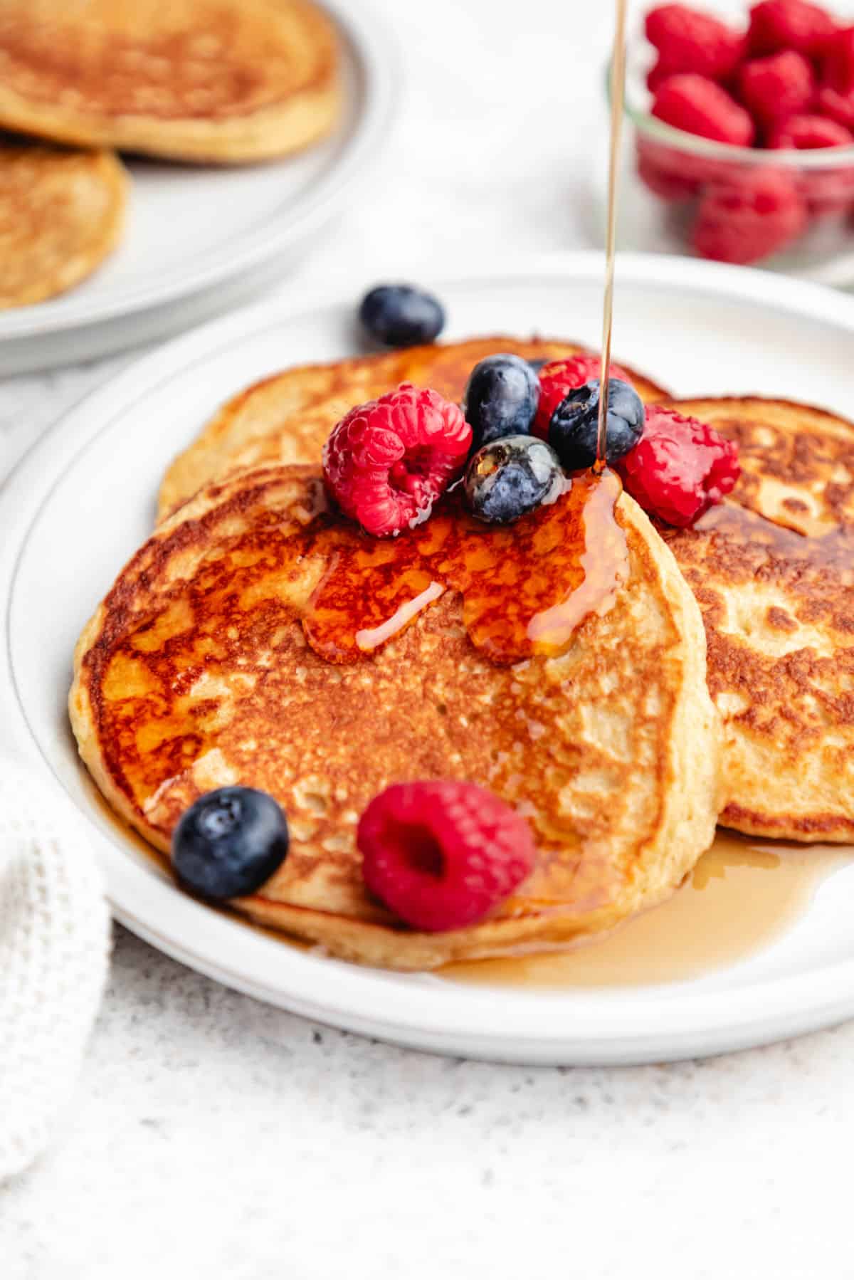 Maple syrup pouring onto cottage cheese pancakes topped with berries.