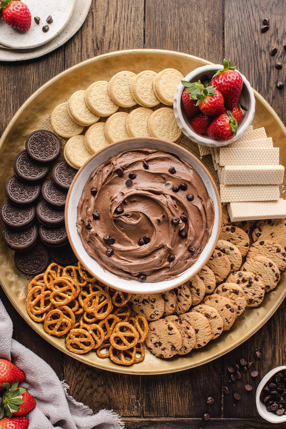 A bowl of brownie batter dip surrounded by strawberries cookies and pretzels.