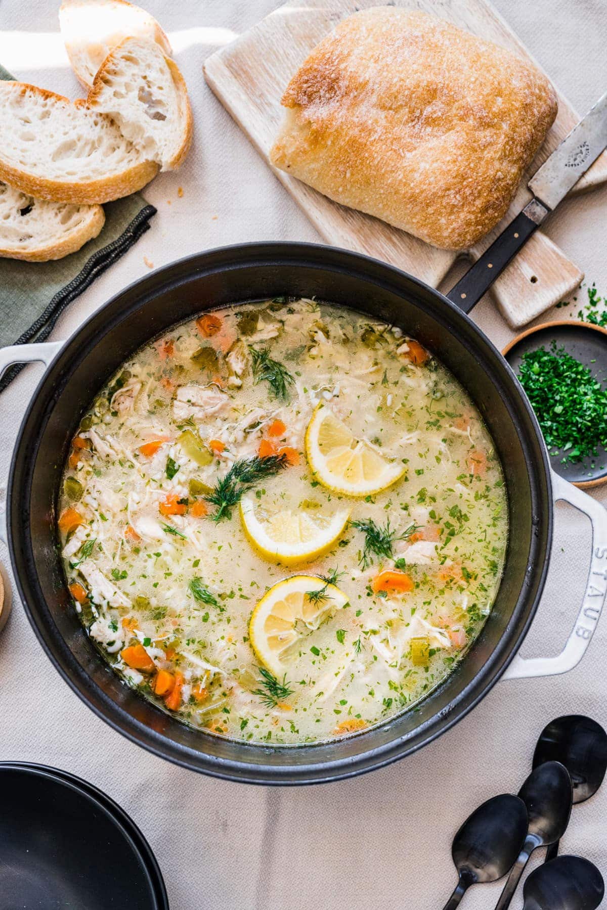 A pot of lemon chicken and rice soup next to bread and spoons.