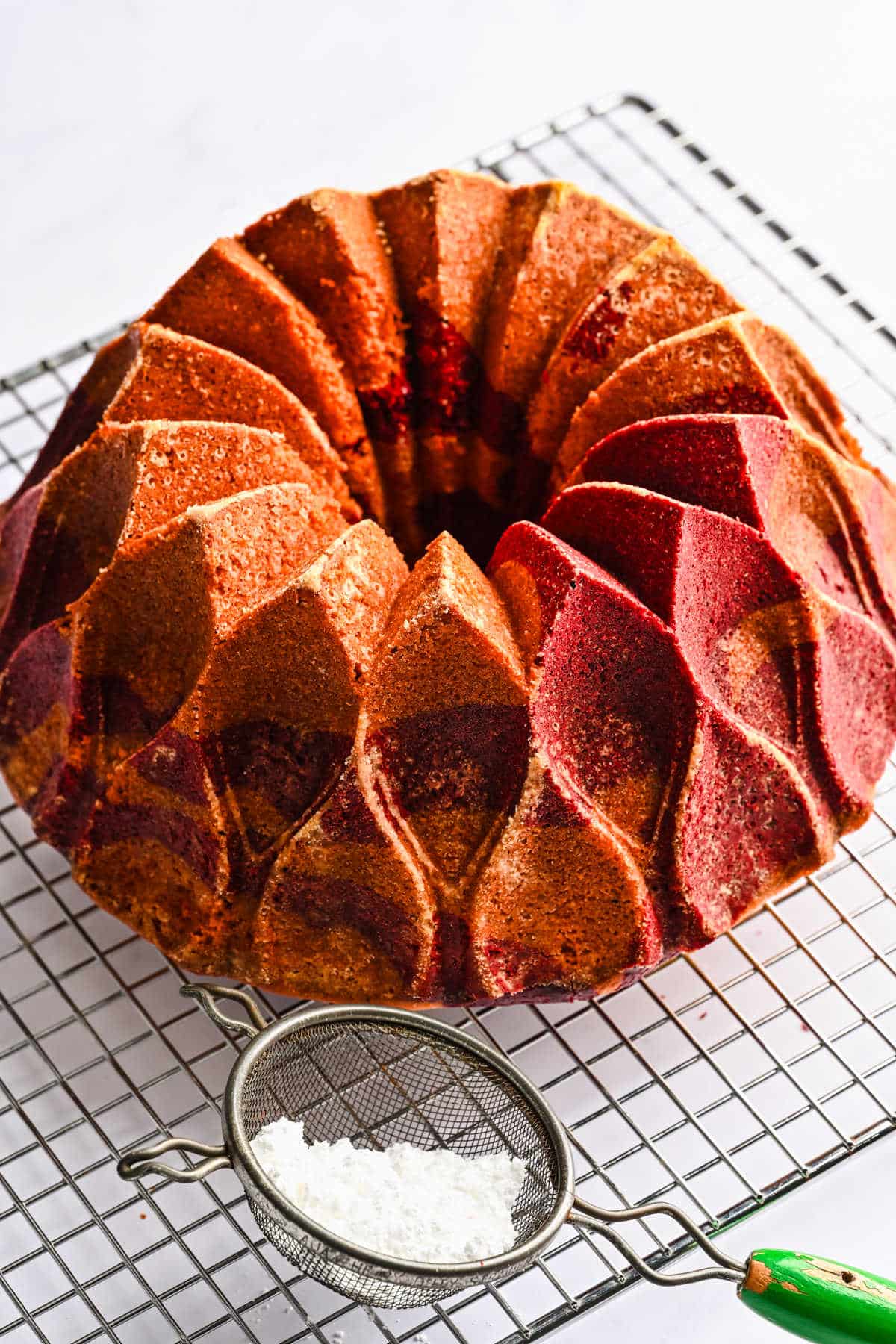 A baked red velvet marble cake on a wire cooling rack next to a powdered sugar sifter. 