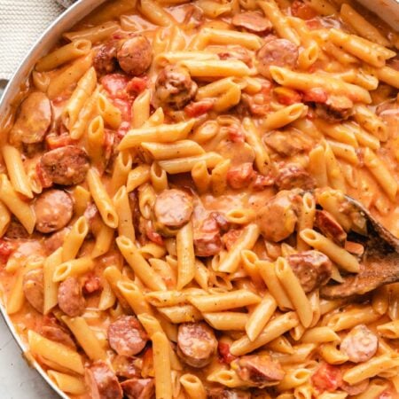 A wooden spoon in a skillet of cheesy kielbasa pasta.