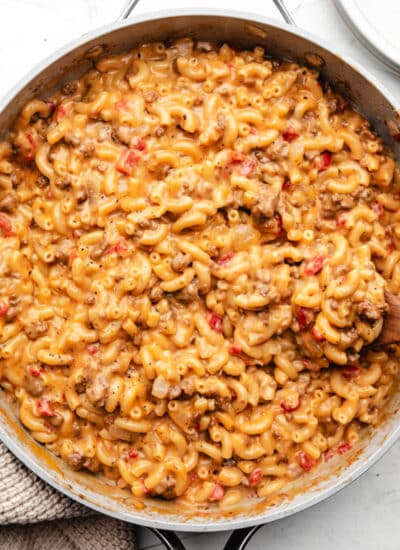 A wooden spoon in a skillet of cheeseburger mac and cheese next to a kitchen cloth.