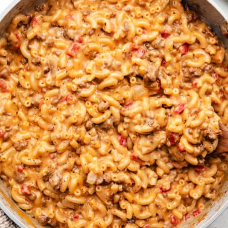 A wooden spoon in a skillet of cheeseburger mac and cheese next to a kitchen cloth.
