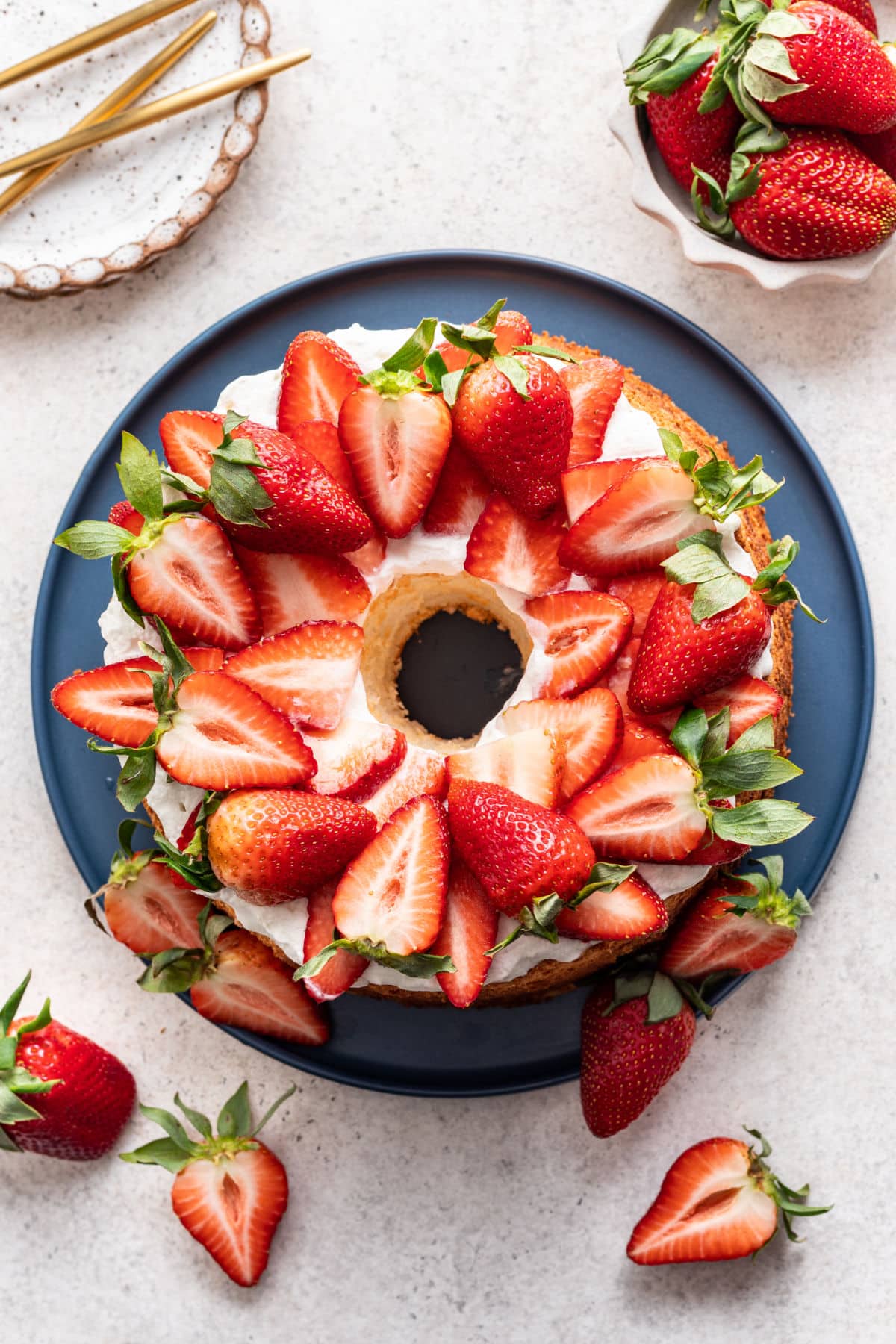 An angel food cake with strawberries and whipped cream on top next to a dish of fresh strawberries. 